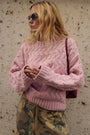Eine Frau mit welligem blondem Haar trägt eine runde Sonnenbrille und einen dicken rosa Pullover mit Zopfmuster. Sie hat eine Camouflage-Hose an und steht vor einer strukturierten Steinmauer, wobei sie eine dunkelrote Umhängetasche bei sich trägt.