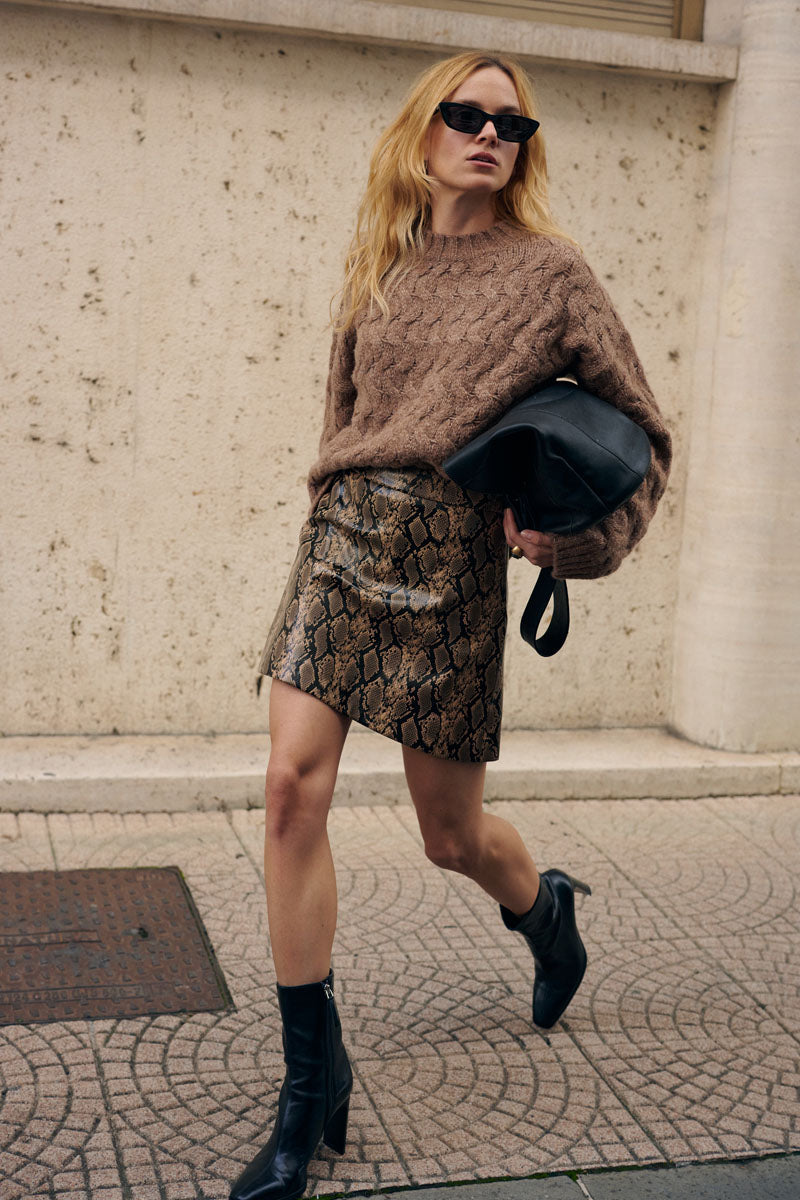 Eine blonde Frau mit dunkler Sonnenbrille geht im Freien spazieren. Sie trägt einen braunen, locker sitzenden Pullover mit Zopfmuster, einen Minirock mit Schlangenhautmuster und schwarze Stiefel mit Absatz. Sie hält eine große schwarze Ledertasche unter dem Arm und lehnt an einer strukturierten beigen Wand.