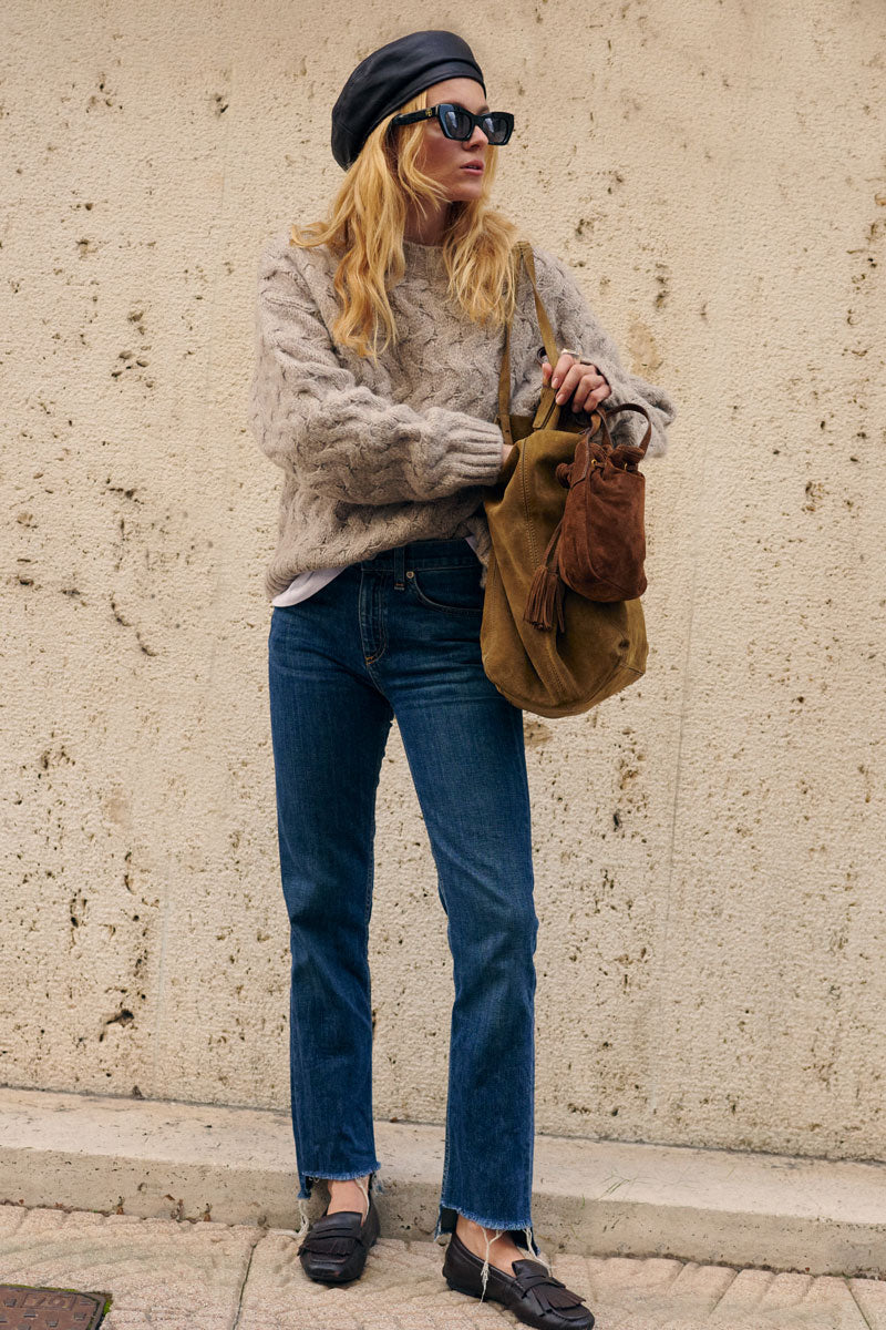 Eine Frau mit langen blonden Haaren trägt eine schwarze Lederbaskenmütze, eine dunkle Sonnenbrille, einen beigen, locker sitzenden Zopfmusterpullover und eine blaue Jeans. Sie steht vor einer strukturierten Steinmauer und trägt eine olivfarbene Umhängetasche aus Wildleder sowie einen braunen Beutel.