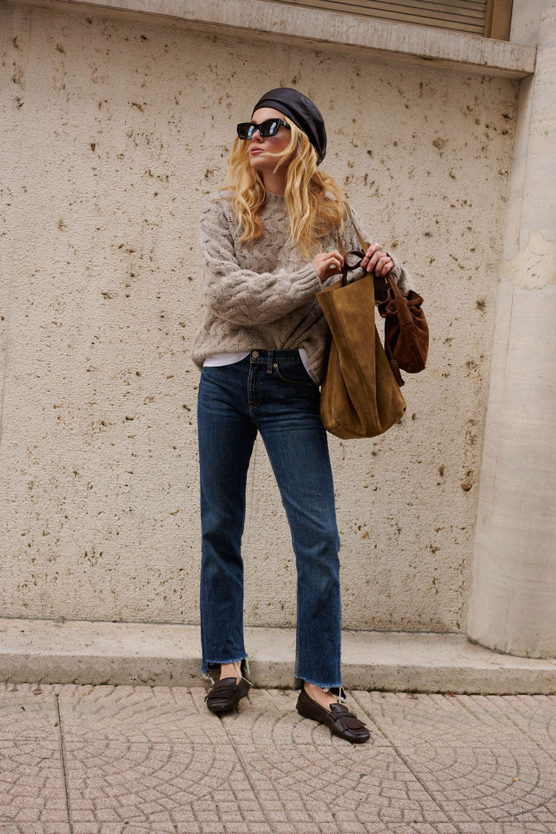 Eine blonde Frau in einem beigen, locker sitzenden Pullover mit Zopfmuster steht vor einer strukturierten Betonwand. Sie trägt eine schwarze Baskenmütze, eine Sonnenbrille, dunkle Jeans und Slipper und hat zwei braune Wildledertaschen dabei.