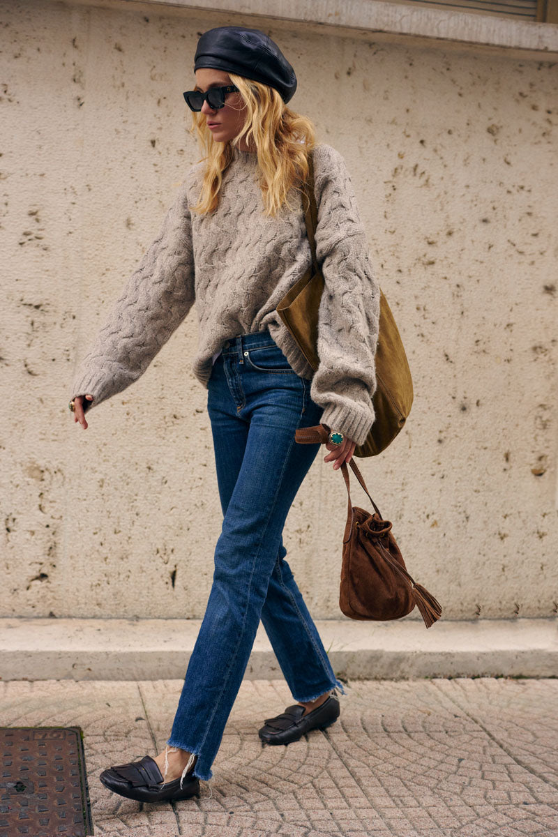 Eine blonde Frau mit schwarzer Baskenmütze, Sonnenbrille und einem locker sitzenden, grob gestrickten Zopfmusterpullover sowie blauen Jeans geht an einer Steinmauer vorbei. Sie trägt eine große olivgrüne Tragetasche und eine braune Wildledertasche sowie schwarze Loafer mit weißen Socken.