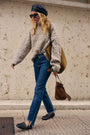 Eine blonde Frau mit schwarzer Baskenmütze, Sonnenbrille und einem locker sitzenden, grob gestrickten Zopfmusterpullover sowie blauen Jeans geht an einer Steinmauer vorbei. Sie trägt eine große olivgrüne Tragetasche und eine braune Wildledertasche sowie schwarze Loafer mit weißen Socken.