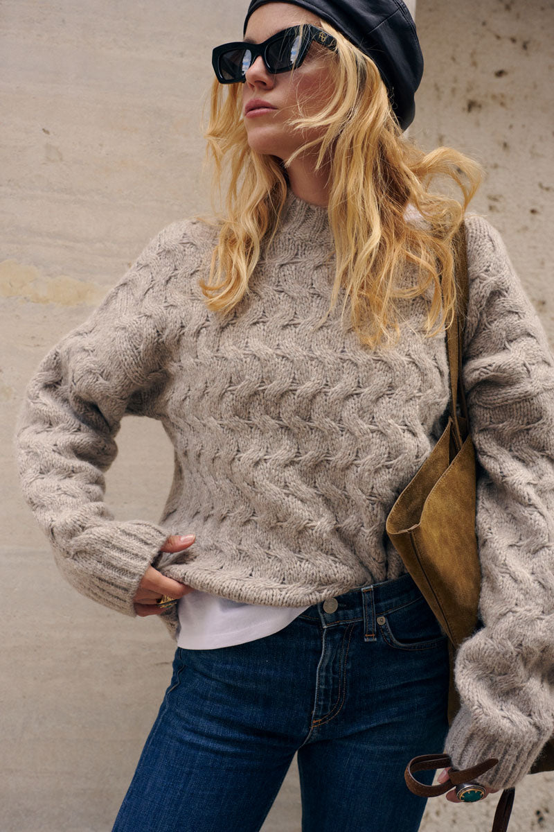 Eine blonde Frau steht vor einer hellen Wand und trägt einen beigen, locker sitzenden Pullover mit Zopfmuster, eine dunkle Jeans und eine schwarze Lederbaskenmütze. Sie trägt eine dunkle Sonnenbrille mit eckigem Rahmen und hat eine olivgrüne Umhängetasche aus Wildleder dabei.