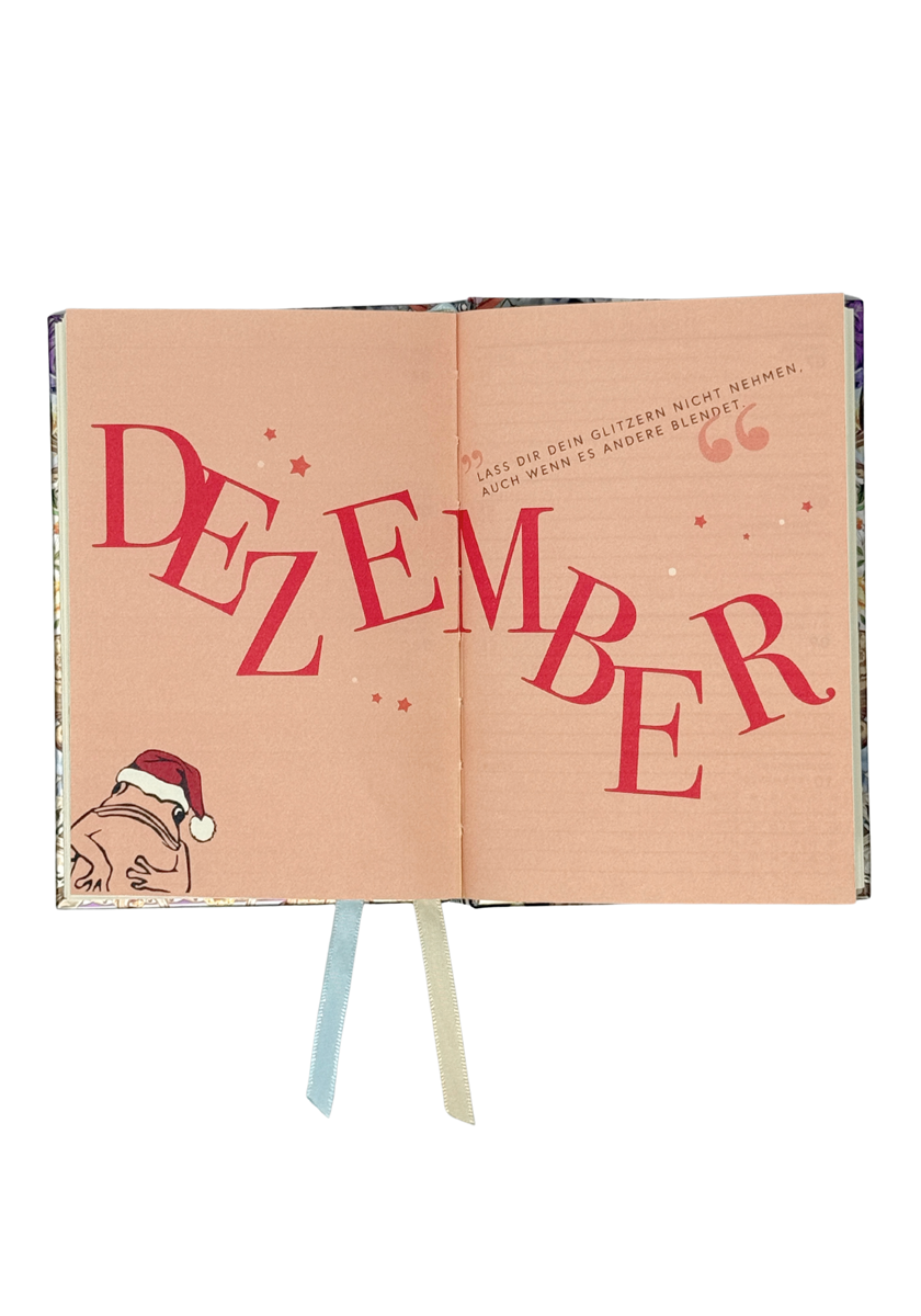 An open Princess Goes Hollywood Jahresplaner 2026 shows peach pages with &quot;DEZEMBER&quot; in bold red, festive stars, a Santa hat-wearing hand on the left, German text on the right page, and two ribbon bookmarks hanging below.