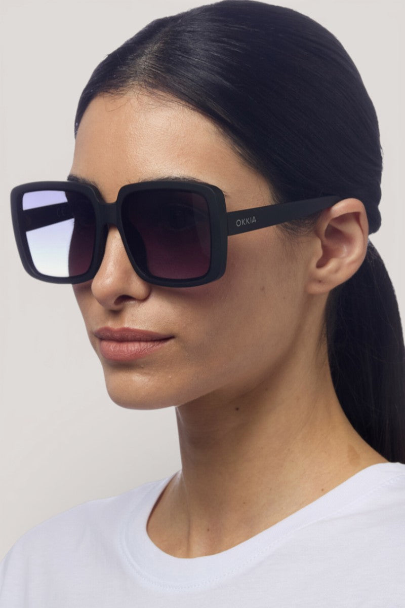 Eine Frau mit glattem dunklem Haar in einem niedrigen Pferdeschwanz trägt Okkia Oversize Sonnenbrille mit einem quadratischen Design, Verlaufsgläsern und UV 400 Schutz. "Okkia" ist auf der Seite aufgedruckt. Sie posiert vor einem schlichten hellen Hintergrund in einem weißen Hemd.