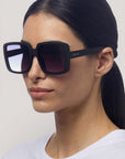 Eine Frau mit glattem dunklem Haar in einem niedrigen Pferdeschwanz trägt Okkia Oversize Sonnenbrille mit einem quadratischen Design, Verlaufsgläsern und UV 400 Schutz. "Okkia" ist auf der Seite aufgedruckt. Sie posiert vor einem schlichten hellen Hintergrund in einem weißen Hemd.