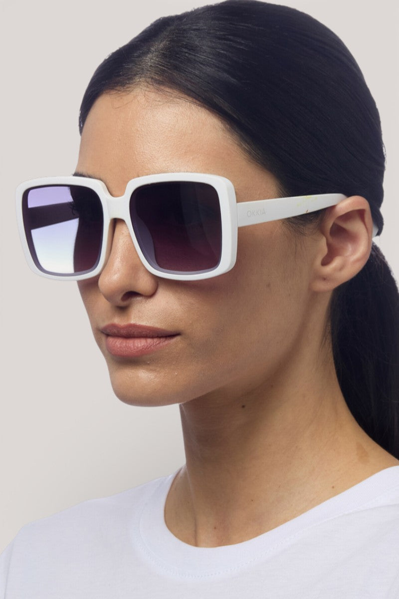 Eine Frau mit glattem, dunklem Haar trägt die übergroße, quadratische Sonnenbrille von Okkia mit dunklen Verlaufsgläsern und UV-400-Schutz. Sie trägt ein schlichtes weißes Oberteil und blickt leicht nach links auf einen hellgrauen Hintergrund.