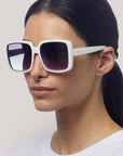 Eine Frau mit glattem, dunklem Haar trägt die übergroße, quadratische Sonnenbrille von Okkia mit dunklen Verlaufsgläsern und UV-400-Schutz. Sie trägt ein schlichtes weißes Oberteil und blickt leicht nach links auf einen hellgrauen Hintergrund.