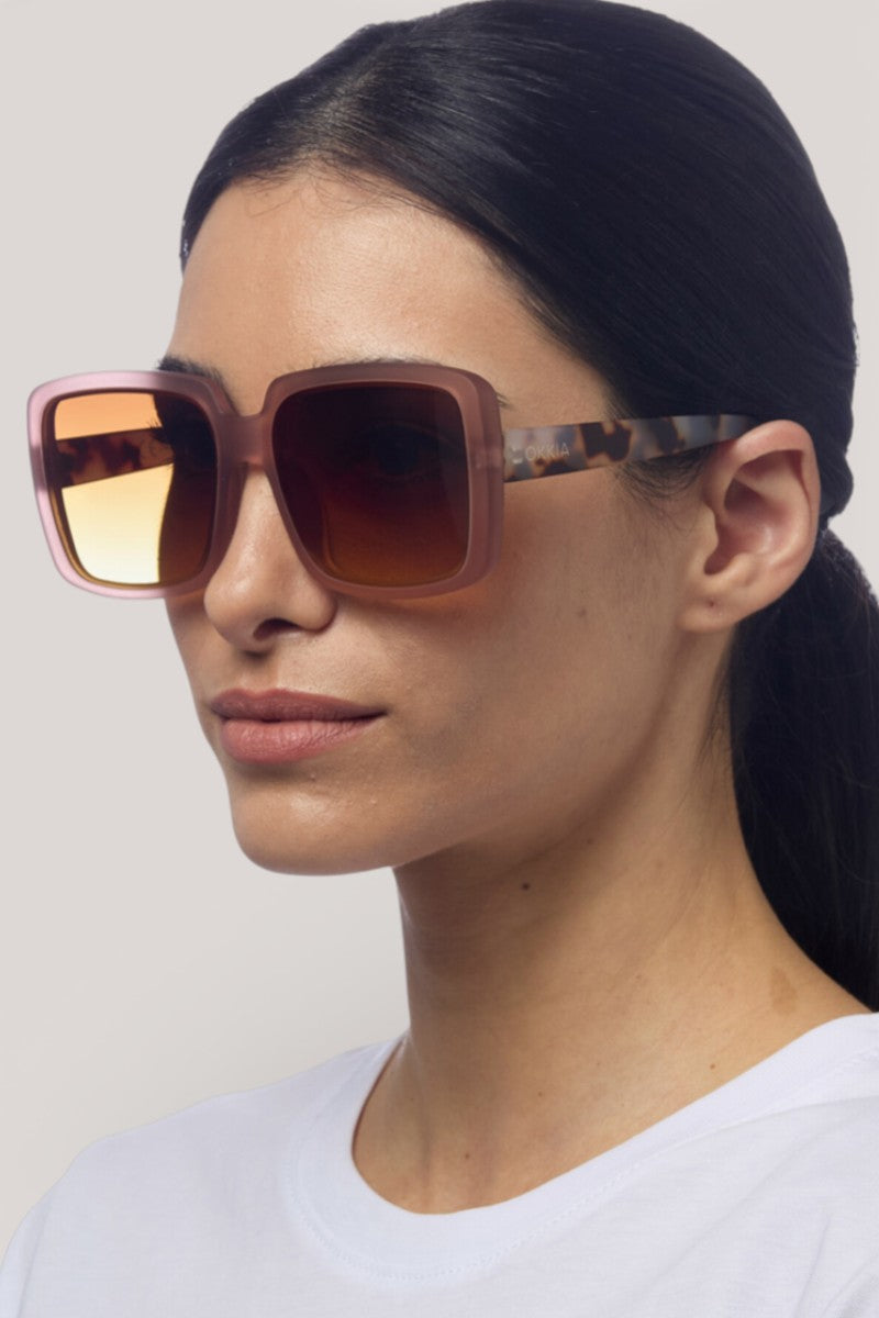 Eine Frau mit glattem, dunklem Haar trägt eine Okkia Oversize Sonnenbrille mit UV 400 Schutz und schildpattgemusterten Bügeln. Die großen quadratischen Rahmen ergänzen ihren neutralen Look und ihr weißes Oberteil vor einem hellgrauen Hintergrund.