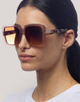 Eine Frau mit glattem, dunklem Haar trägt eine Okkia Oversize Sonnenbrille mit UV 400 Schutz und schildpattgemusterten Bügeln. Die großen quadratischen Rahmen ergänzen ihren neutralen Look und ihr weißes Oberteil vor einem hellgrauen Hintergrund.