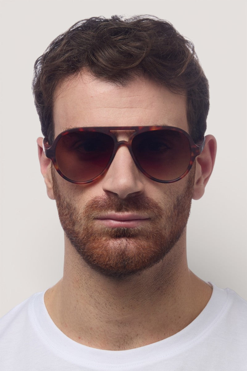 Ein Mann mit kurzen, gewellten braunen Haaren und einem rötlich-braunen Bart trägt eine große, unisex Okkia Aviator Sonnenbrille mit UV 400-Schutz und ein weißes T-Shirt mit Rundhalsausschnitt. Er blickt nach vorne und hat einen neutralen Gesichtsausdruck vor einem schlichten hellen Hintergrund.
