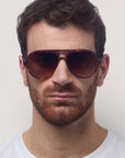 Ein Mann mit kurzen, gewellten braunen Haaren und einem rötlich-braunen Bart trägt eine große, unisex Okkia Aviator Sonnenbrille mit UV 400-Schutz und ein weißes T-Shirt mit Rundhalsausschnitt. Er blickt nach vorne und hat einen neutralen Gesichtsausdruck vor einem schlichten hellen Hintergrund.