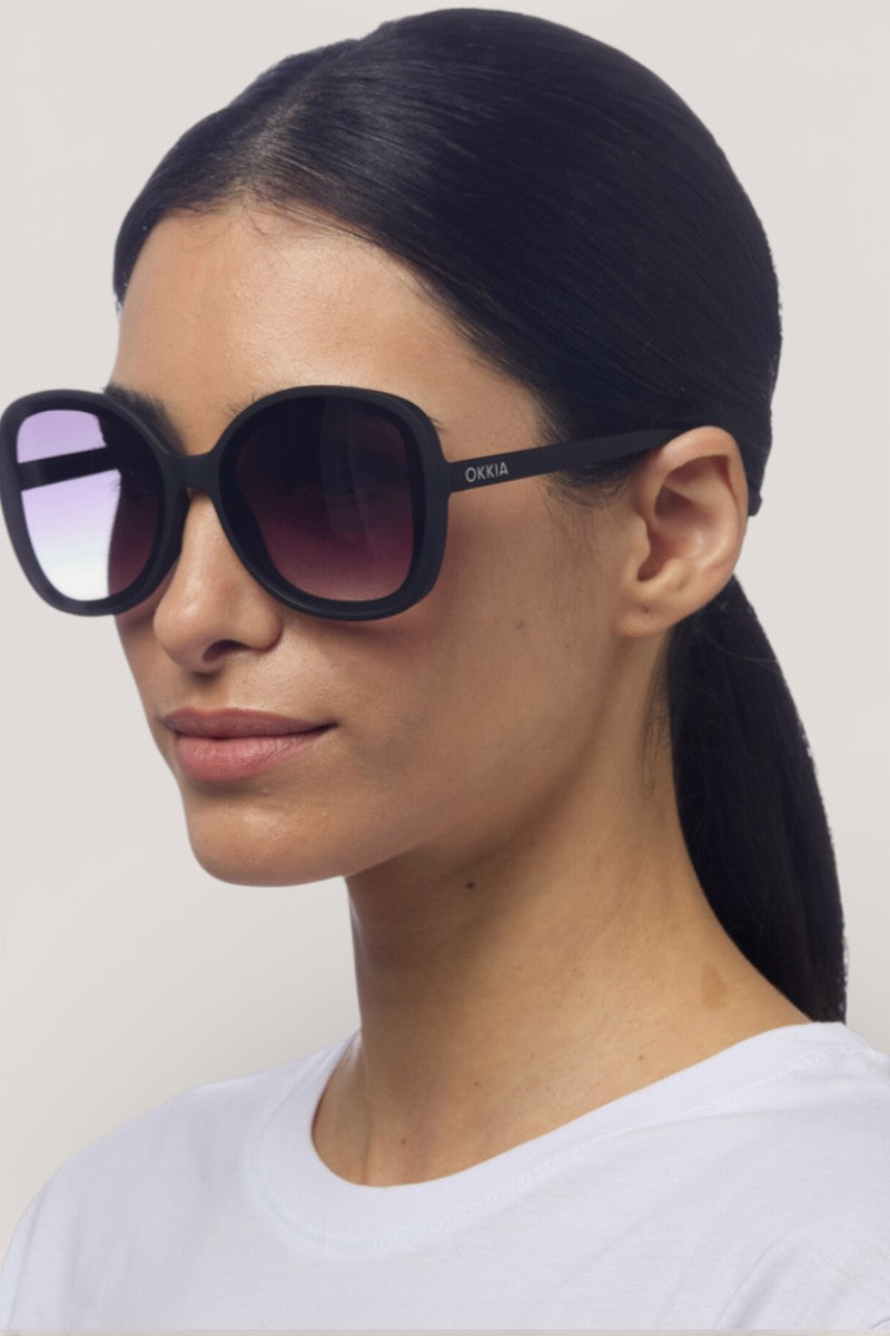 Eine Frau mit glattem schwarzem Haar in einem niedrigen Pferdeschwanz trägt eine große, runde Okkia-Sonnenbrille mit UV-400-Schutz und Verlaufsgläsern. Ihr neutraler Gesichtsausdruck und ihr weißes Hemd betonen die auffälligen Okkia-Fassungen vor einem schlichten hellen Hintergrund.