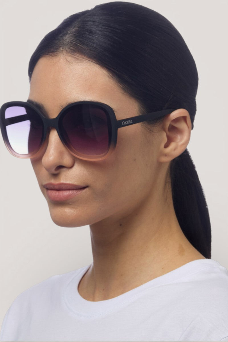 Eine Frau mit langen, nach hinten gebundenen dunklen Haaren trägt eine übergroße Okkia ANNA-Sonnenbrille mit schickem Schmetterlingsdesign, dezentem Branding und UV-400-Schutz. Sie hat einen neutralen Gesichtsausdruck, ein weißes Oberteil und steht vor einem schlichten hellen Hintergrund.