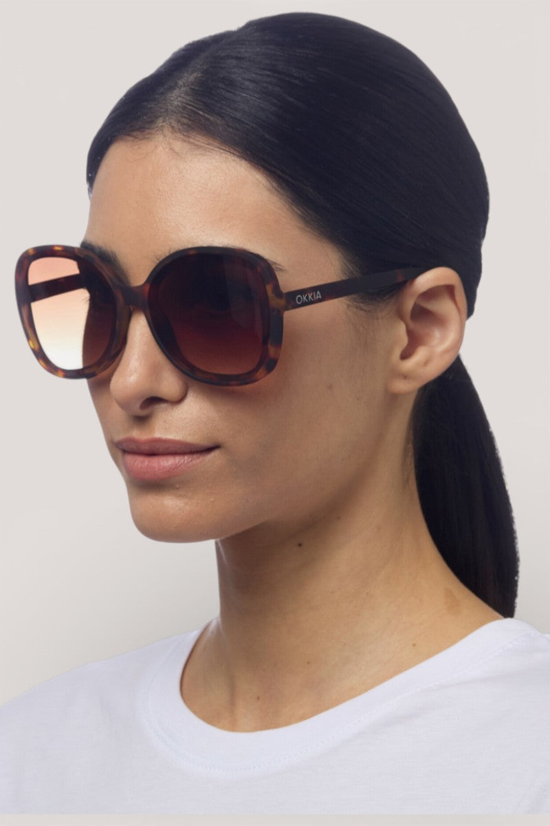 Eine Frau mit glattem, dunklem Haar in einem niedrigen Pferdeschwanz trägt eine große, runde, schildpattfarbene Okkia-Sonnenbrille mit Verlaufsgläsern und UV-400-Schutz. Sie posiert mit einem weißen Hemd vor einem schlichten hellen Hintergrund.