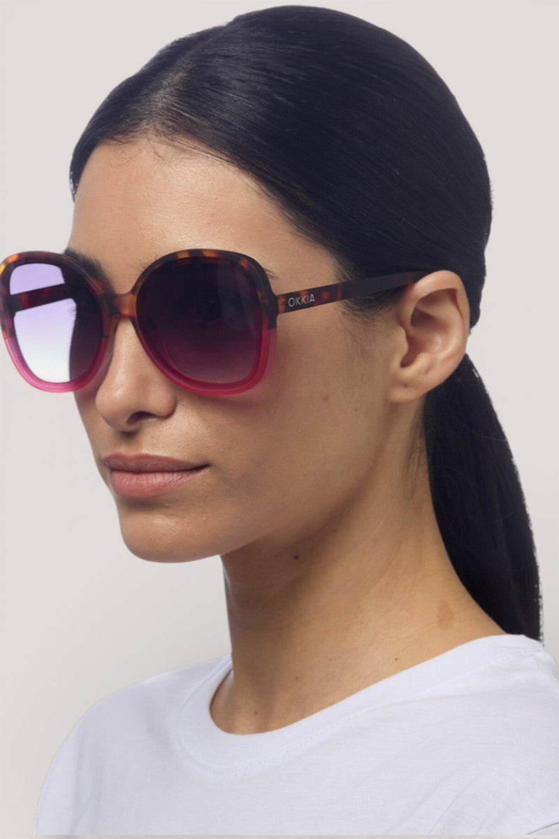 Eine Frau mit glattem, nach hinten gebundenem dunklem Haar trägt eine große, runde Sonnenbrille von Okkia Sonnenbrille mit schildpattfarbenem Rahmen und rosa Gläsern. Sie hat einen neutralen Gesichtsausdruck, trägt ein schlichtes weißes Hemd und steht vor einem hellgrauen Hintergrund.