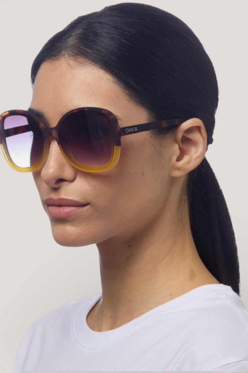 Eine Frau mit glattem dunklem Haar in einem niedrigen Pferdeschwanz trägt die ANNA Sonnenbrille von Okkia - eine große, runde Sonnenbrille mit braunem und gelbem Rahmen und UV-400-Schutz. Sie trägt ein schlichtes weißes Oberteil vor einem hellgrauen Hintergrund.