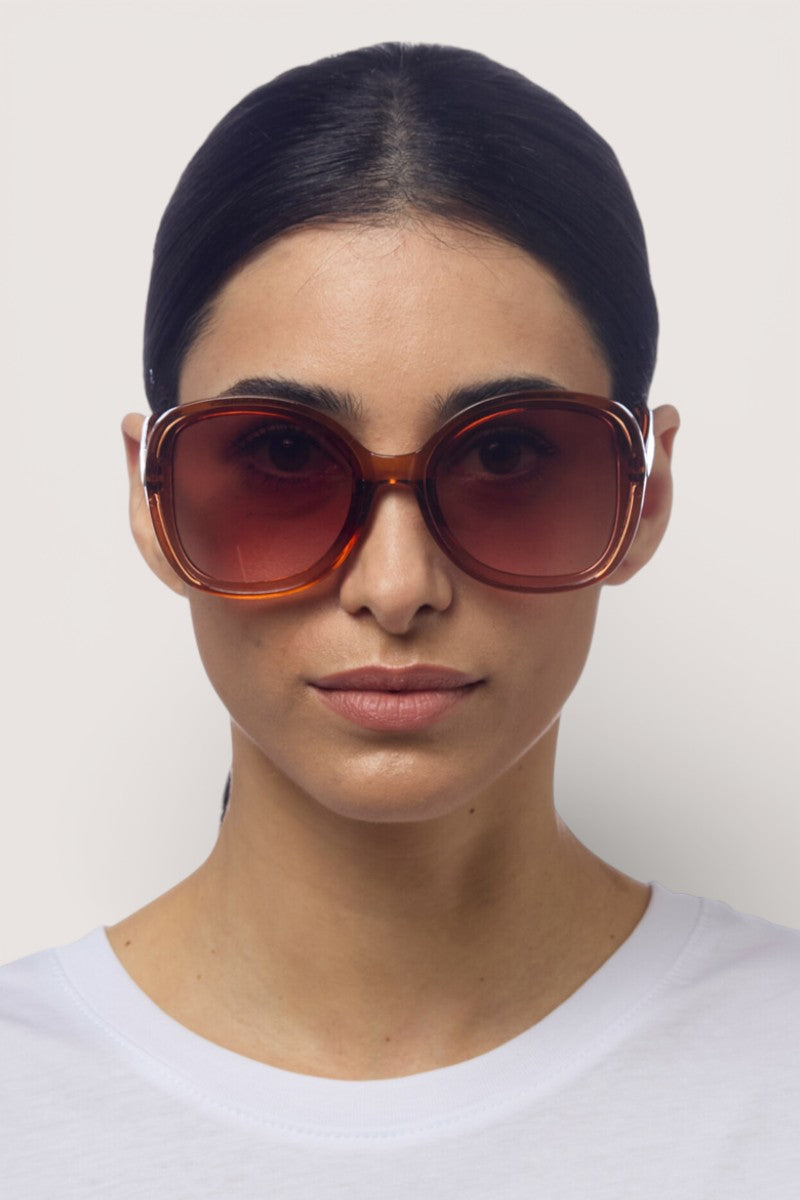 Eine Frau mit glattem, dunklem Haar trägt eine übergroße Okkia-Sonnenbrille mit UV-400-Schutz und ein weißes Rundhalsshirt mit Schmetterlingsmotiv. Sie blickt nach vorne vor einem schlichten hellen Hintergrund, ihr Gesichtsausdruck ist neutral.