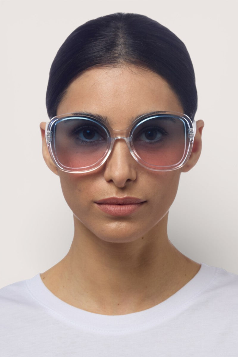 Eine Frau mit dunklem, zurückgegeltem Haar trägt eine übergroße, runde Okkia-Sonnenbrille mit rosa getönten Gläsern und UV-400-Schutz. Sie hat einen neutralen Gesichtsausdruck und trägt ein schlichtes weißes Hemd vor einem hellen, neutralen Hintergrund.