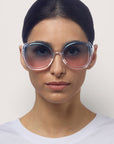 Eine Frau mit dunklem, zurückgegeltem Haar trägt eine übergroße, runde Okkia-Sonnenbrille mit rosa getönten Gläsern und UV-400-Schutz. Sie hat einen neutralen Gesichtsausdruck und trägt ein schlichtes weißes Hemd vor einem hellen, neutralen Hintergrund.