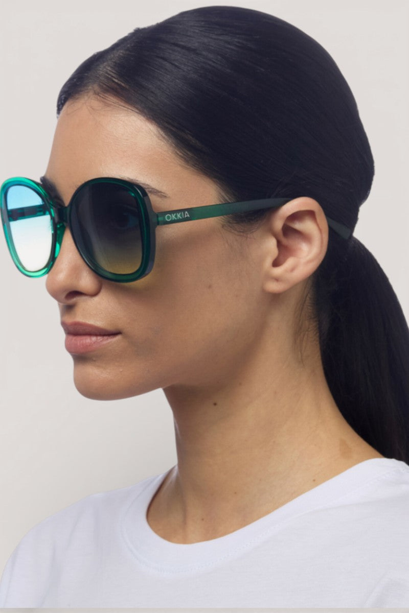 Eine Frau mit glattem schwarzem Haar in einem niedrigen Pferdeschwanz trägt eine große, runde grüne Okkia Sonnenbrille mit UV-400-Schutz und Verlaufsgläsern. Sie hat einen neutralen Gesichtsausdruck und ist mit einem schlichten weißen Hemd vor einem einfachen hellen Hintergrund bekleidet.