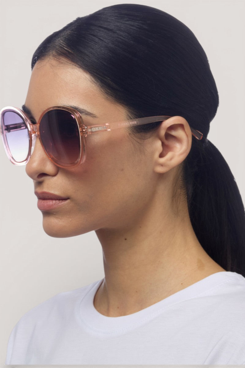 Frau mit glattem dunklem Haar in einem niedrigen Pferdeschwanz trägt eine runde rosafarbene Sonnenbrille im Butterfly-Design (UV-400-Schutz) von Okkia und ein weißes T-Shirt mit Rundhalsausschnitt, dargestellt im Profil vor einem schlichten hellen Hintergrund.