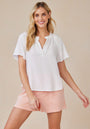 A woman with long blonde hair smiles in a Bella Dahl V-neck 100% Lyocell Bluse and light pink shorts. She stands relaxed against a beige background, one hand at her side, the other slightly outstretched.