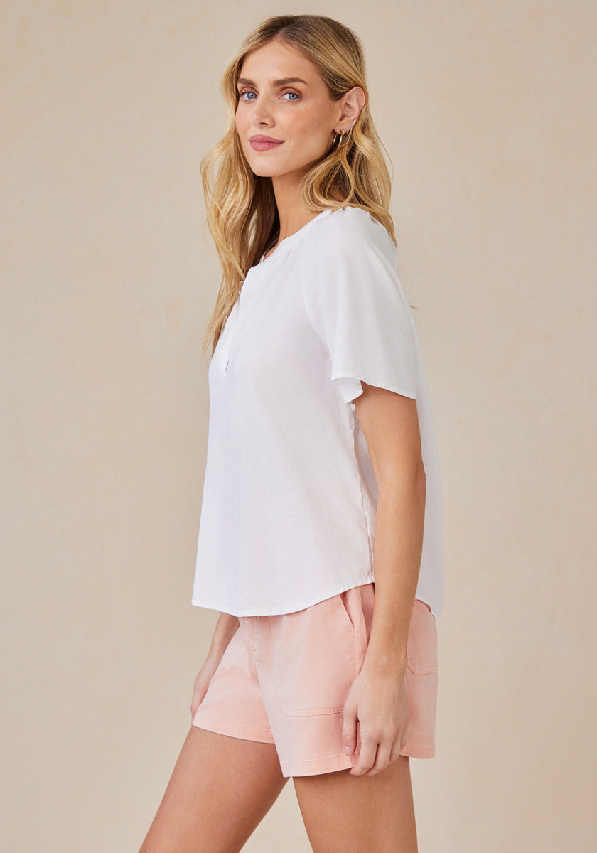 A woman with long blonde hair stands sideways, wearing a Bella Dahl locker geschnittene Bluse mit V-Ausschnitt and light pink shorts. She smiles slightly and looks forward against a plain beige background.