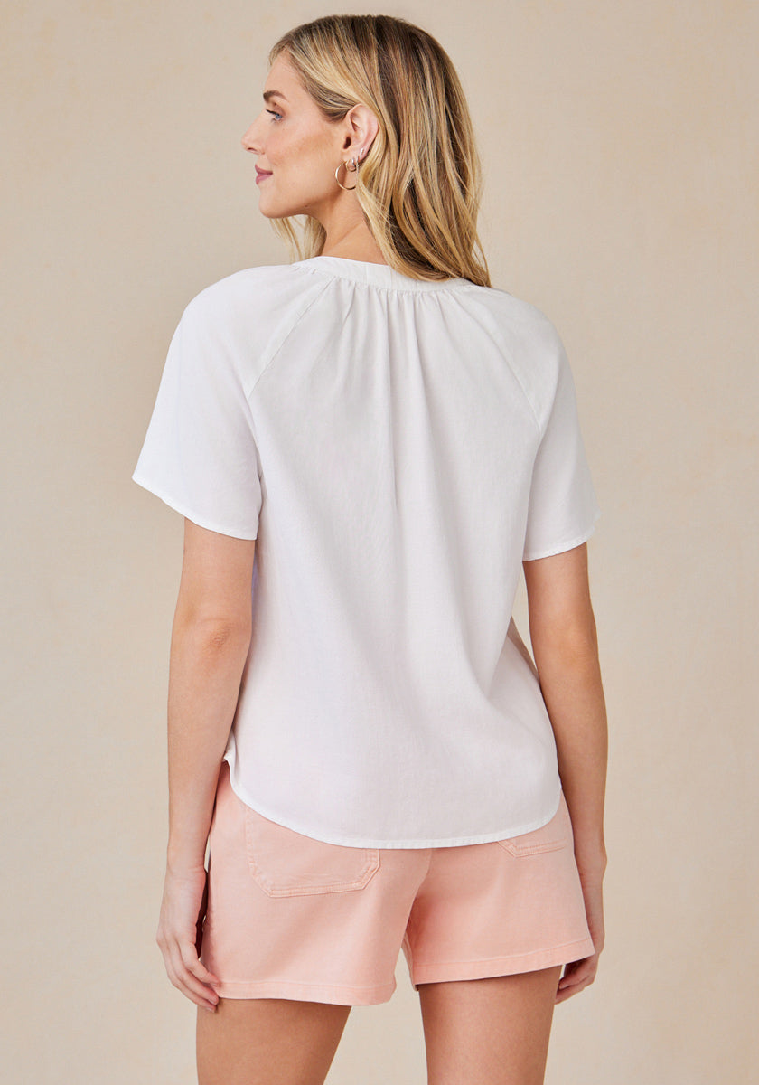 A woman faces away, displaying the back of her Bella Dahl locker geschnittene Lyocell-Bluse with V-Ausschnitt, paired with light pink shorts. Her straight blonde hair falls down against a neutral beige background.