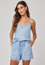 A woman with long wavy brown hair wears a light blue Bella Dahl Lyocell sleeveless top with a frayed v-neckline and matching drawstring shorts in denim look, standing against a plain background with her hands in her pockets, smiling gently.