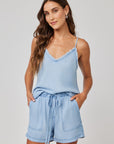 A woman with long wavy brown hair wears a light blue Bella Dahl Lyocell sleeveless top with a frayed V-neck and matching drawstring shorts with an elastic waistband. She stands hands in pockets, smiling softly at the camera against a plain background.