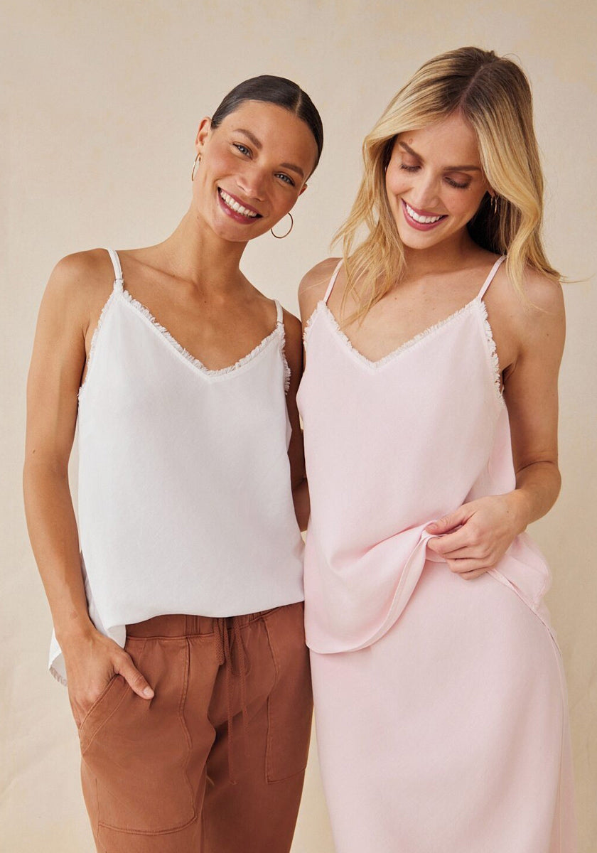 Two women smile together. The left wears a white Bella Dahl Lyocell sleeveless top with brown pants, her hair in a low bun and hand in pocket. The right, blonde with hair down, wears a light pink Bella Dahl V-Ausschnitt camisole and matching skirt.