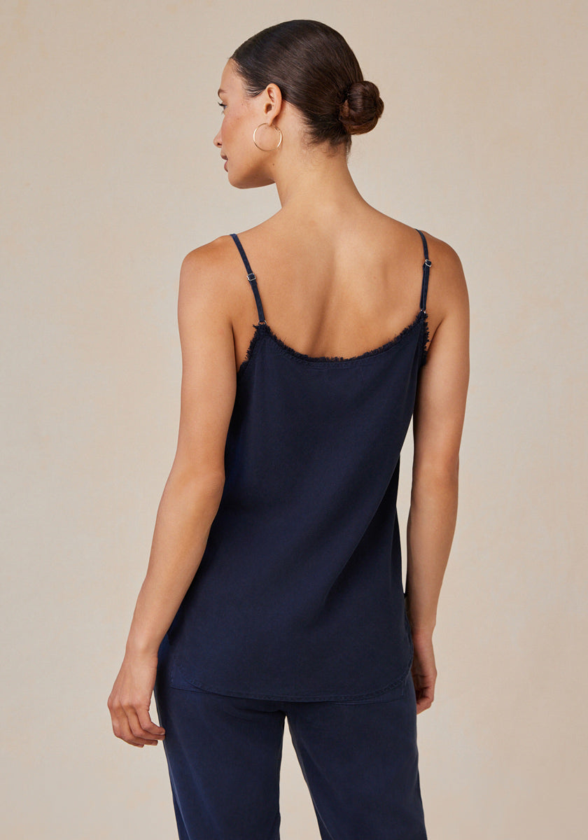 A woman with medium skin tone and brown hair in a neat bun, seen from the back, wears a Bella Dahl navy blue V-neck sleeveless camisole with thin straps and matching denim-look pants, standing against a light beige background.