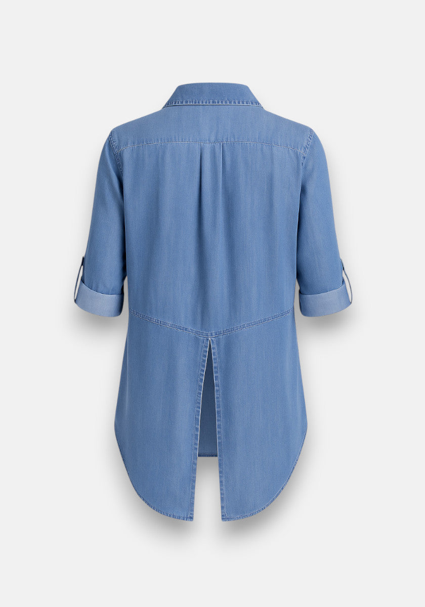 Bella Dahl’s light blue denim shirt in 100% Lyocell is shown from the back, with rolled-up sleeves and buttoned tabs, pleated upper back yoke, center back hem slit, Hemdkragen collar, and a rounded high-low hemline.