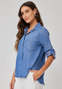 A woman with long wavy brown hair wears a light blue Bella Dahl Denimoptik shirt with Hemdkragen, made from 100% Lyocell. She stands against a plain background, smiling softly with one hand in her pocket and looking slightly left.
