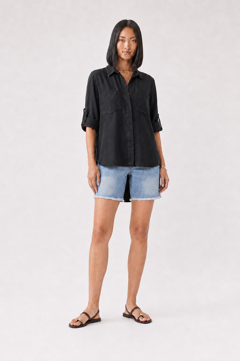 Eine Frau mit glattem, schulterlangem schwarzem Haar trägt ein locker sitzendes „Bella Dahl Damen Hemd“ in Schwarz, dazu hellblaue Jeansshorts mit einem Schlitz vorne und schwarze Sandalen. Sie steht mit dem Blick nach vorne vor einem hellen, neutralen Hintergrund.