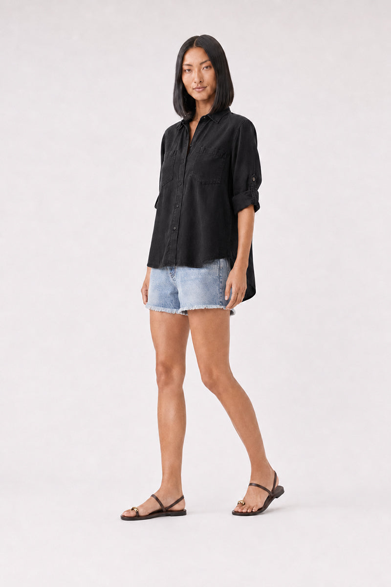 Eine Frau mit glattem schwarzem Haar trägt ein locker sitzendes schwarzes Bella Dahl Damenhemd, hellblaue, ausgefranste Jeansshorts und schwarze Sandalen. Sie steht vor einem schlichten hellgrauen Hintergrund, blickt mit entspanntem Gesichtsausdruck nach vorne und hält die Arme an den Seiten.