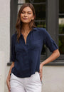 A woman with long brown hair stands outdoors in front of a white wall and black-trimmed window, wearing a navy blue Bella Dahl Denimoptik Hemd aus 100% Lyocell with rolled-up sleeves and Brusttaschen, paired with white pants.