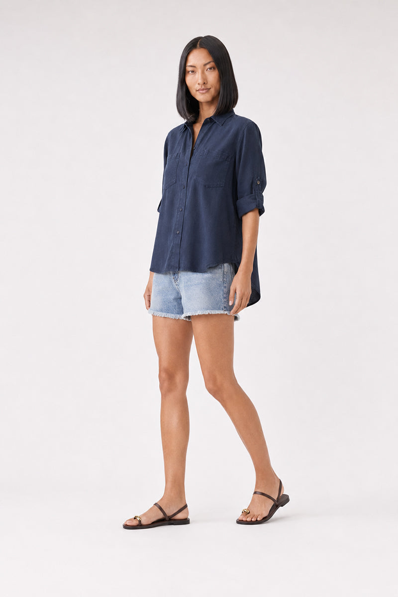 Eine Person mit glattem, schulterlangem schwarzem Haar trägt ein locker sitzendes „Bella Dahl Lyocell Hemd“ in Marineblau mit hochgekrempelten Ärmeln, hellblaue Jeansshorts und schwarze flache Sandalen. Sie steht vor einem schlichten hellen Hintergrund und blickt mit neutralem Gesichtsausdruck in die Kamera.