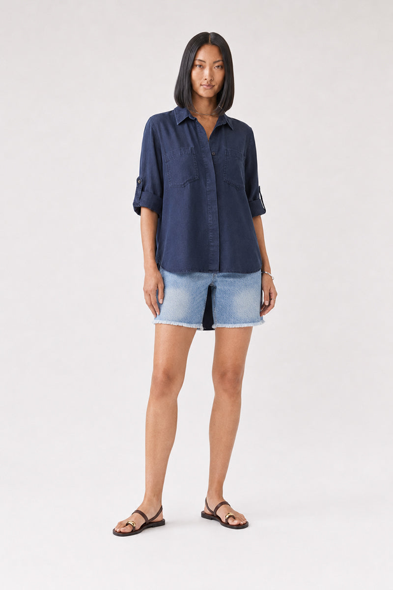 Eine Frau mit glattem, dunklem Haar trägt ein marineblaues Lyocell-Hemd von Bella Dahl mit hochgekrempelten Ärmeln und hellblaue, ausgefranste Jeansshorts. Sie steht mit dem Blick nach vorne, die Hände entspannt an den Seiten, und trägt schwarze Sandalen vor einem schlichten, hellen Hintergrund.