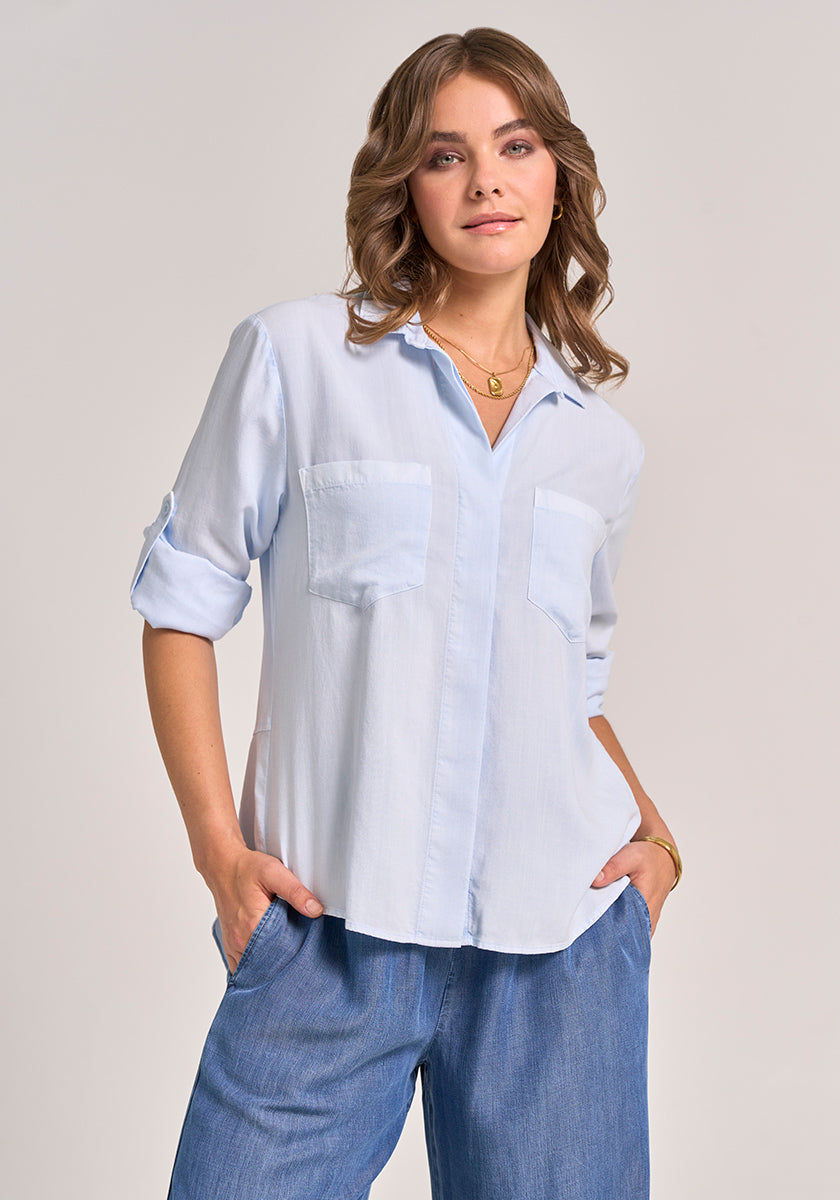 A woman with wavy light brown hair stands against a plain background, smiling gently as she wears gold jewelry and a Bella Dahl light blue button-up shirt with classic collar, two front pockets, and denim-look lyocell fabric.