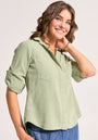 A woman with wavy brown hair smiles while wearing a Bella Dahl sage green Lyocell button-up shirt featuring rolled sleeves, chest pockets, and a Knopfleiste. She pairs it with blue jeans against a plain, light background. One hand rests near her face.