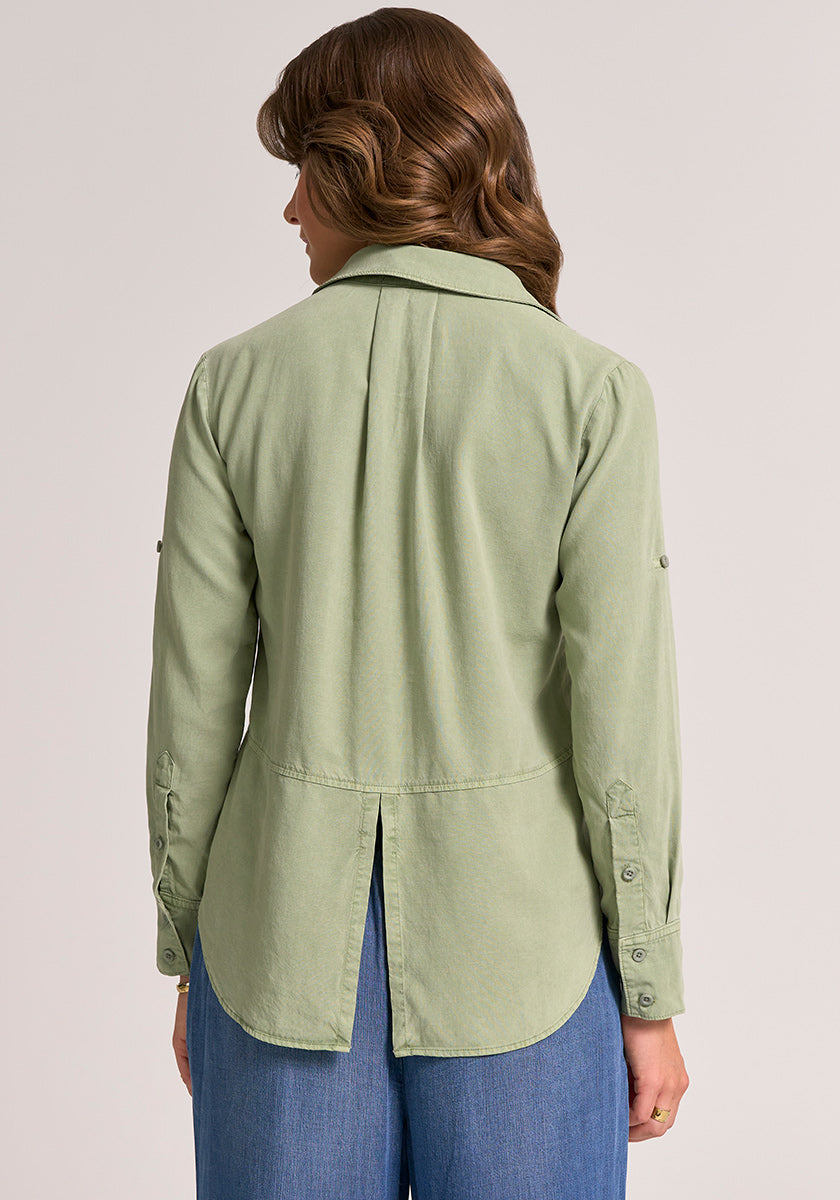 A woman with wavy brown hair, seen from behind, wears a light green long-sleeve Lyocell button-up shirt by Bella Dahl with Knopfleiste, back vent, and curved hem, paired with blue wide-leg pants against a plain light background.