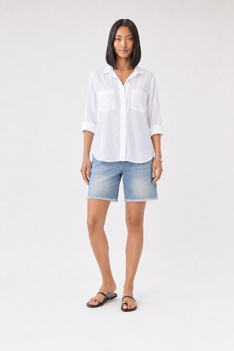 Vor einem schlichten Hintergrund steht eine Frau, die ein weißes Bella Dahl Damenhemd mit hochgekrempelten Ärmeln, hellblaue Jeansshorts, schwarze Sandalen und ein Armband trägt. Sie hat glattes, schulterlanges schwarzes Haar und einen neutralen Gesichtsausdruck; ihre Arme liegen entspannt an den Seiten.