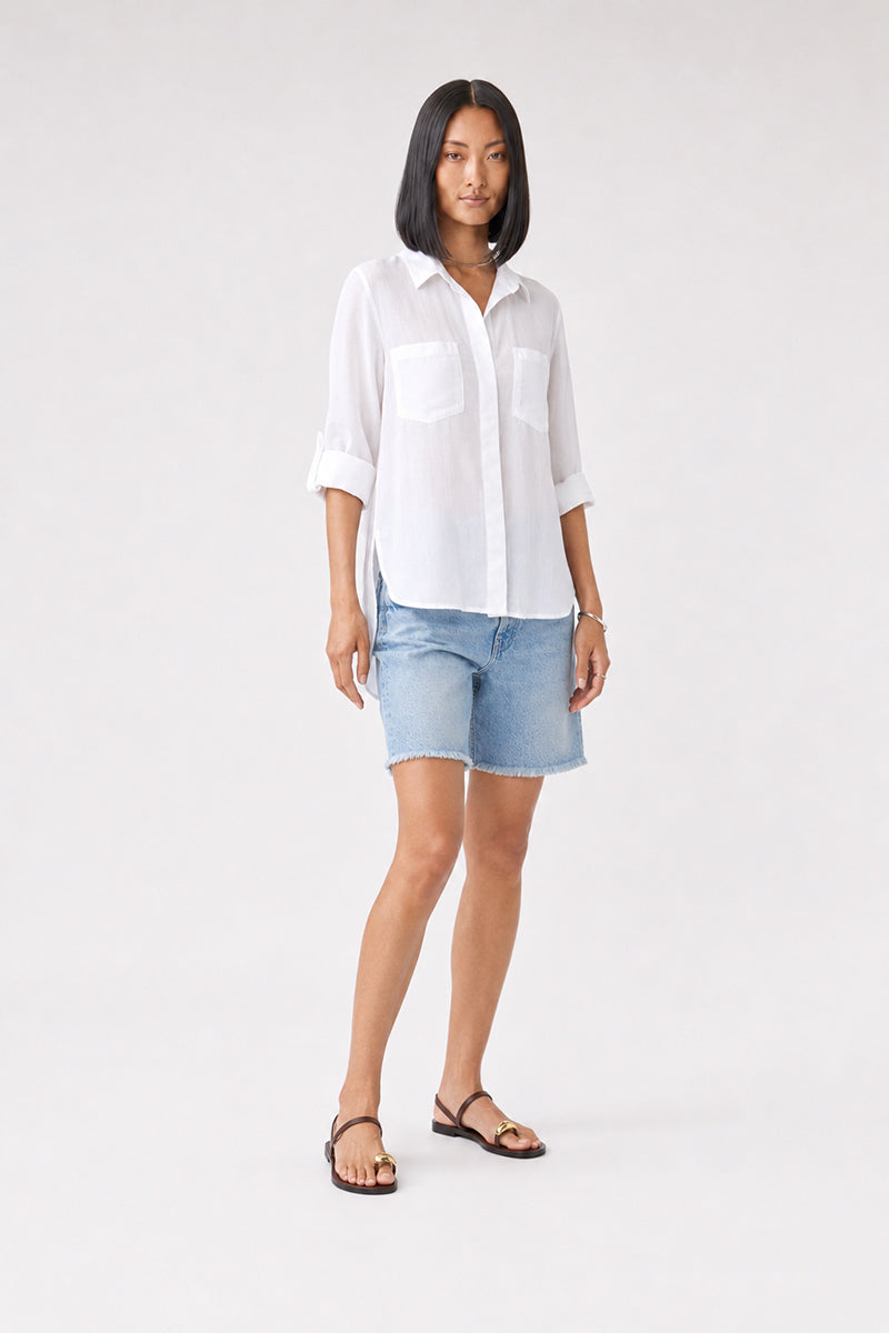 Eine Frau steht mit dem Blick nach vorne und trägt ein durchsichtiges weißes Bella Dahl Damenhemd mit hochgekrempelten Ärmeln, hellblaue, knielange Jeansshorts, schwarze Sandalen und eine Halskette. Ihr Haar ist glatt und schulterlang. Der Hintergrund ist einfarbig hellgrau.