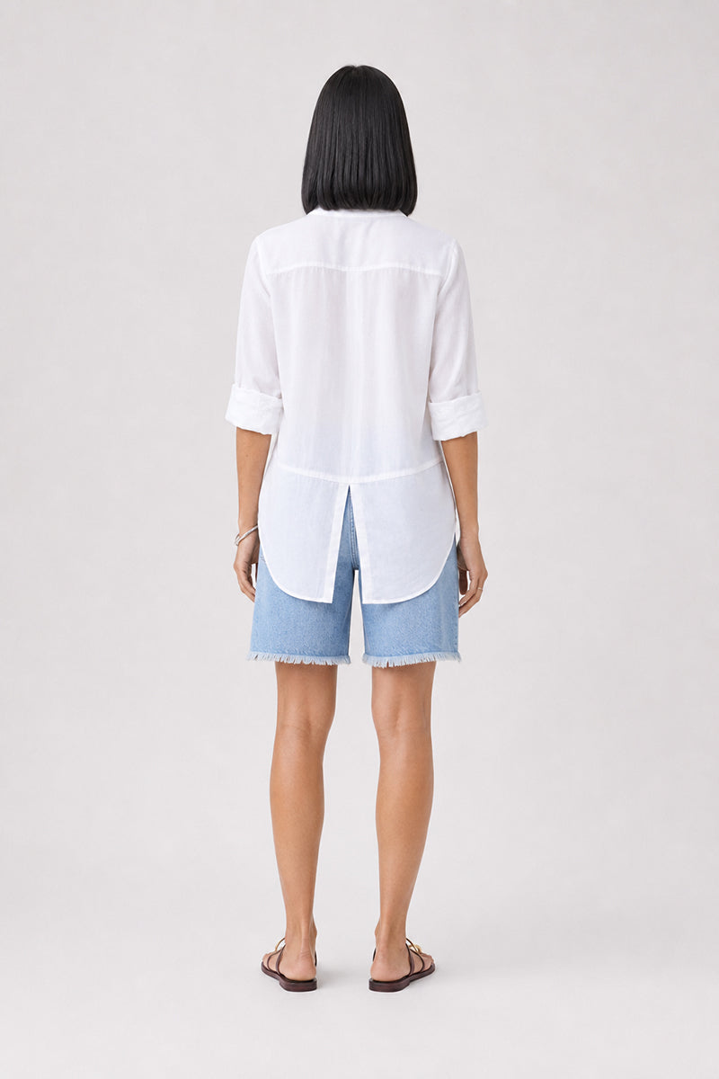 Eine Person steht mit dem Rücken zur Kamera und trägt ein weißes Bella Dahl Damenhemd mit umgeschlagenen Ärmelbündchen, blaue Jeansshorts mit ausgefransten Säumen und braune Sandalen. Sie hat glattes, schwarzes, schulterlanges Haar und posiert vor einem schlichten hellgrauen Hintergrund.