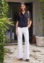 A woman stands on a stone patio wearing a Bella Dahl short-sleeved black shirt with Knopfleiste, wide-leg white pants, black sandals, and carrying a woven tan shoulder bag. Green vines and a planter decorate the doorway behind her.