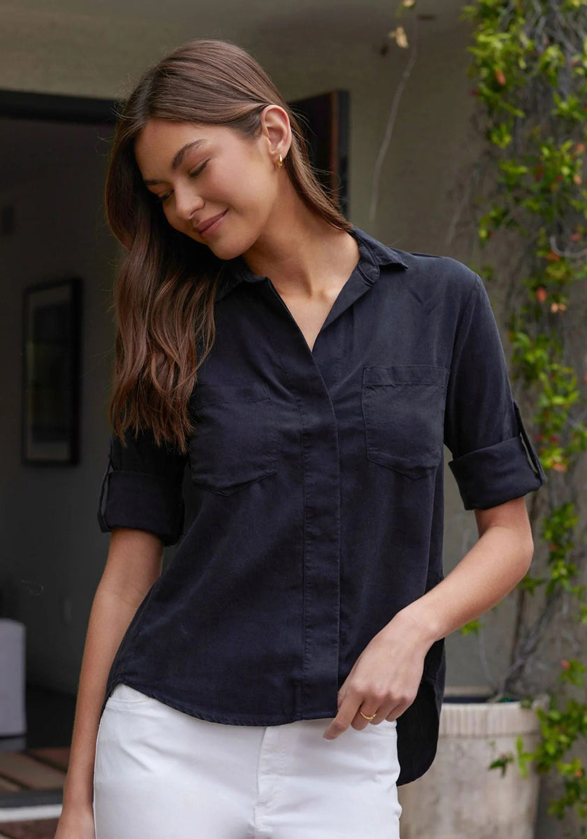 A woman with long brown hair stands outside, smiling softly. She wears a black Bella Dahl Lyocell Hemd with rolled sleeves and white pants. Greenery and a modern building are visible in the background.