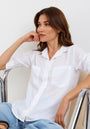 A woman with light skin and brown hair sits on a metal chair, resting her chin on her hand. She wears a loose Bella Dahl Lyocell Hemd with rolled sleeves and blue Jeans in Denimoptik, gazing thoughtfully against a white background.
