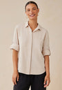 A woman with medium skin tone and brown hair in a ponytail smiles at the camera. She wears gold hoop earrings, black pants, and a beige Bella Dahl Lyocell button-up shirt with Hemdkragen and rolled sleeves against a light beige background.